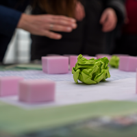 Tisch mit einem Projektplan und verschiedenen rosa Würfeln. In der Mitte ein zerknüllter grüner Zettel. Im Hintergrund sind die Hände von Personen zu sehen, die über dem Tisch etwas zeigen.