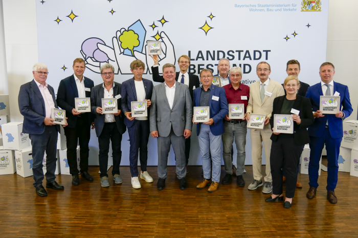 Die Vertreter der elf Modellgemeinden im Projekt Landstadt Bestand und Staatsminister Christian Bernreiter stehen vor einer Pressewand.