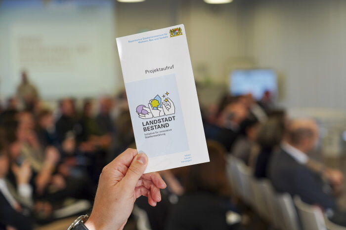 Ein Hand hält den Flyer zum Projektaufruf des neuen Modellvorhabens LANDSTADT BESTAND in Höhe.