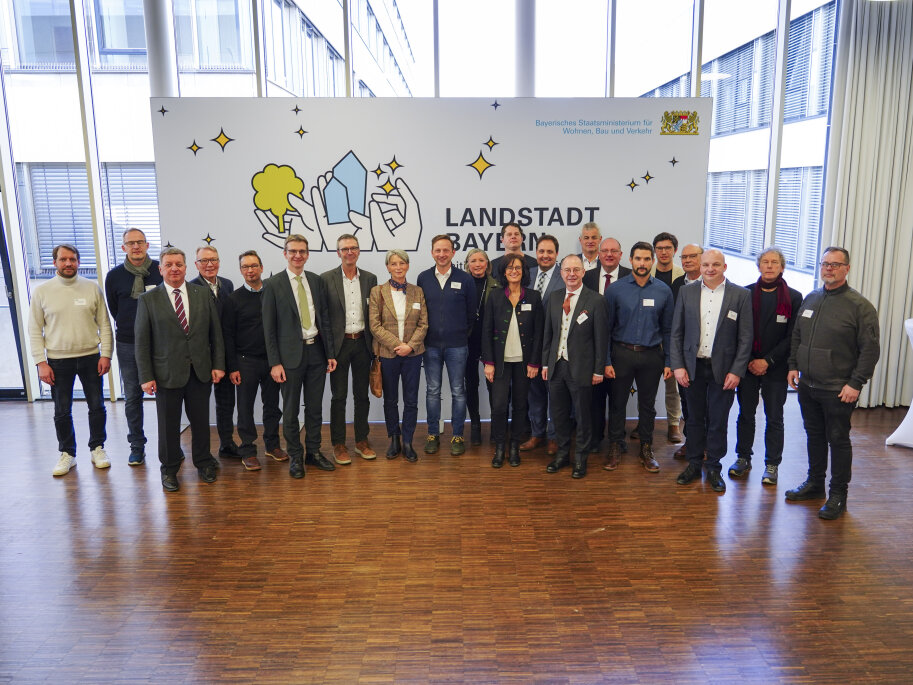Alle Vertreterinnen und Vertreter der zehn Modellgemeinden und Staatsminister Christian Bernreiter stehen vor der Pressewand LANDSTADT BAYERN.